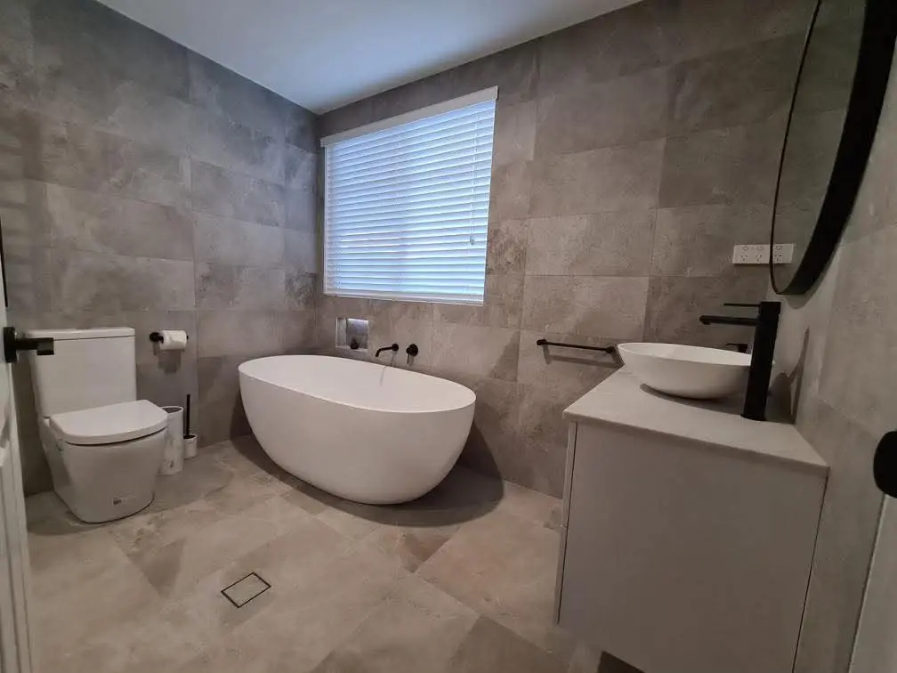 Stylish bathroom showcasing a sleek white freestanding bathtub with bathtub sealing & waterproofing, complemented by tiled walls, a modern sink, toilet, and a bright window with blinds.