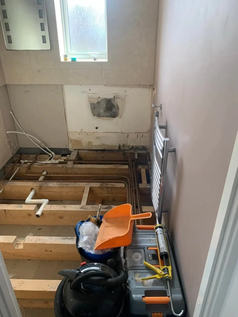A bathroom under renovation for a leaks repair service, showing exposed wooden flooring, visible plumbing pipes, and a damaged wall, with tools like a caulking gun and a shop vac ready for use, alongside a towel radiator and a small window.