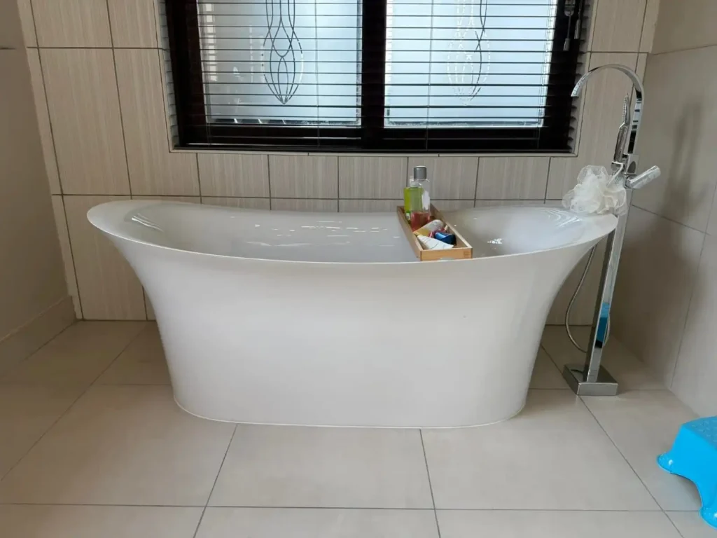 A modern bathroom featuring a new freestanding white bathtub, highlighting the need for an Old Bathtub Removal Service to replace an outdated fixture. The space includes tiled walls, a large window with blinds, a wooden tray with bath products, a chrome faucet, and a blue step stool on the tiled floor.