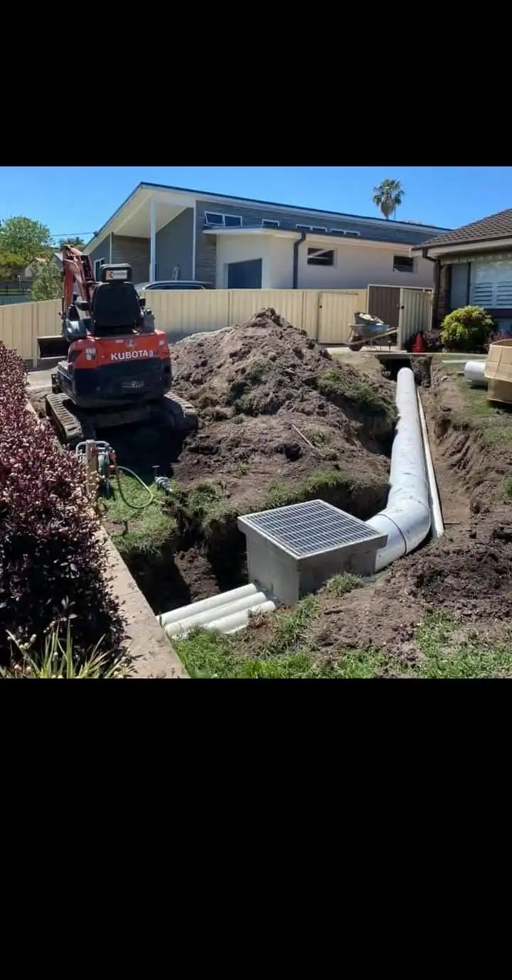 A construction site for plumbing and drainage upgrades, featuring a mini excavator digging a trench with pipes and a drainage grate, near a residential house.