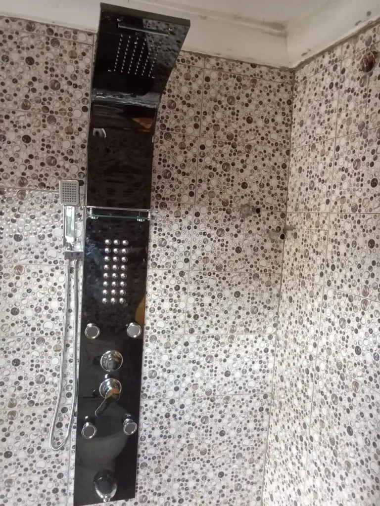 A sleek shower panel with a modern black finish, featuring a large overhead showerhead, multiple adjustable jets, and a handheld shower with a flexible hose, set against a stylish bathroom wall with patterned tiles.