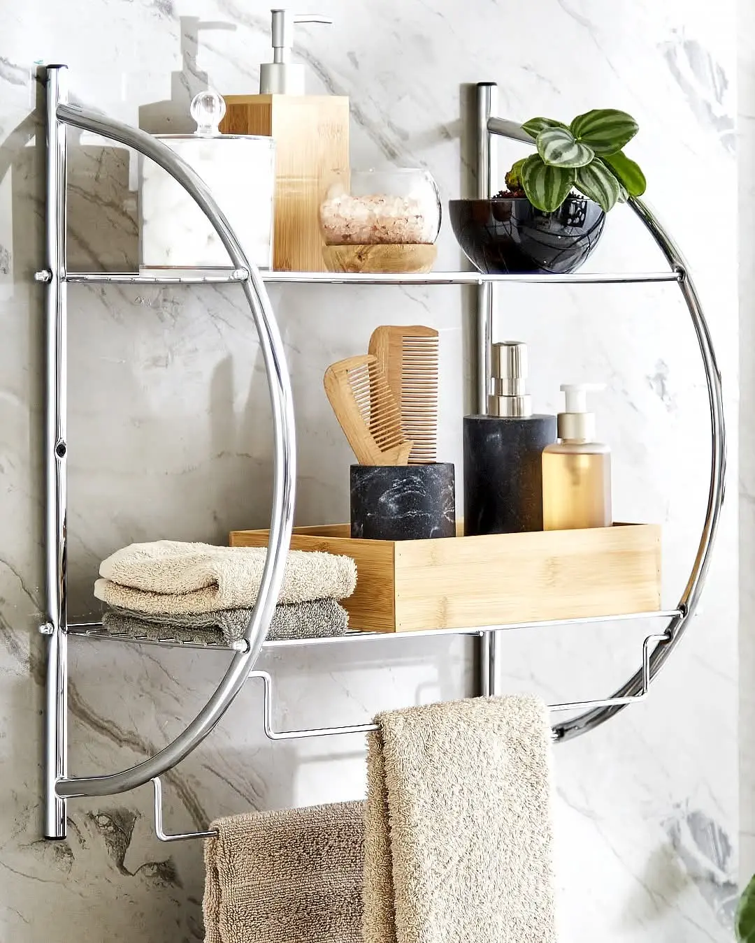 A sleek small storage solution with a curved chrome shelf, holding towels, toiletries, a plant, and a bamboo box, mounted on a stylish marble wall.