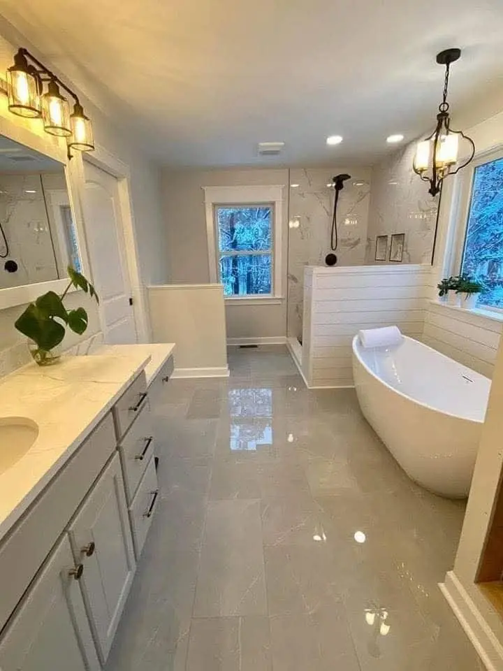 A stylish bathroom featuring a modern Bathtub Installation with a freestanding white bathtub as the focal point. The room includes a double vanity with a marble countertop, a large mirror, and pendant lighting. The space also has a glass-enclosed shower, tiled walls, and glossy gray flooring, with natural light coming through a window.