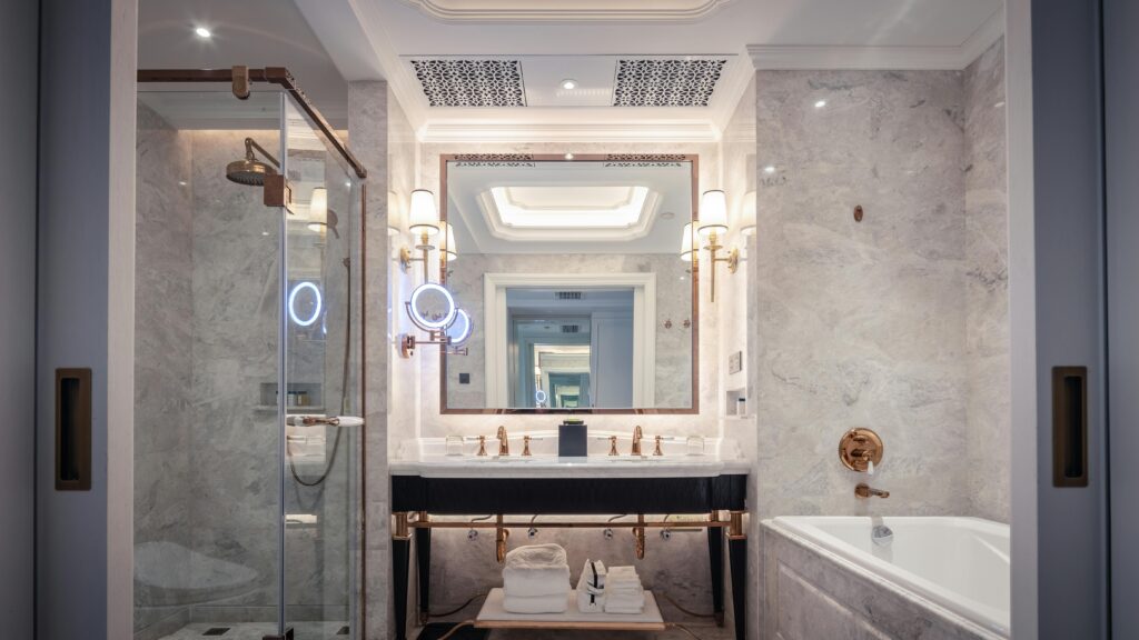 A modern, elegant bathroom featuring marble walls, a glass-enclosed walk-in shower on the left, and a bathtub on the right. A double-sink vanity with gold fixtures sits beneath a large mirror with wall-mounted lights, creating a bright, luxurious atmosphere.
